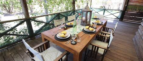 Una terraza de madera con una mesa dispuesta para comer, rodeada de sillas y una vista a follaje a través de una barandilla de madera.
