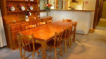 A wooden dining table with six chairs, a kitchen area with a stove and cabinets, and a shelf with plates and flowers.