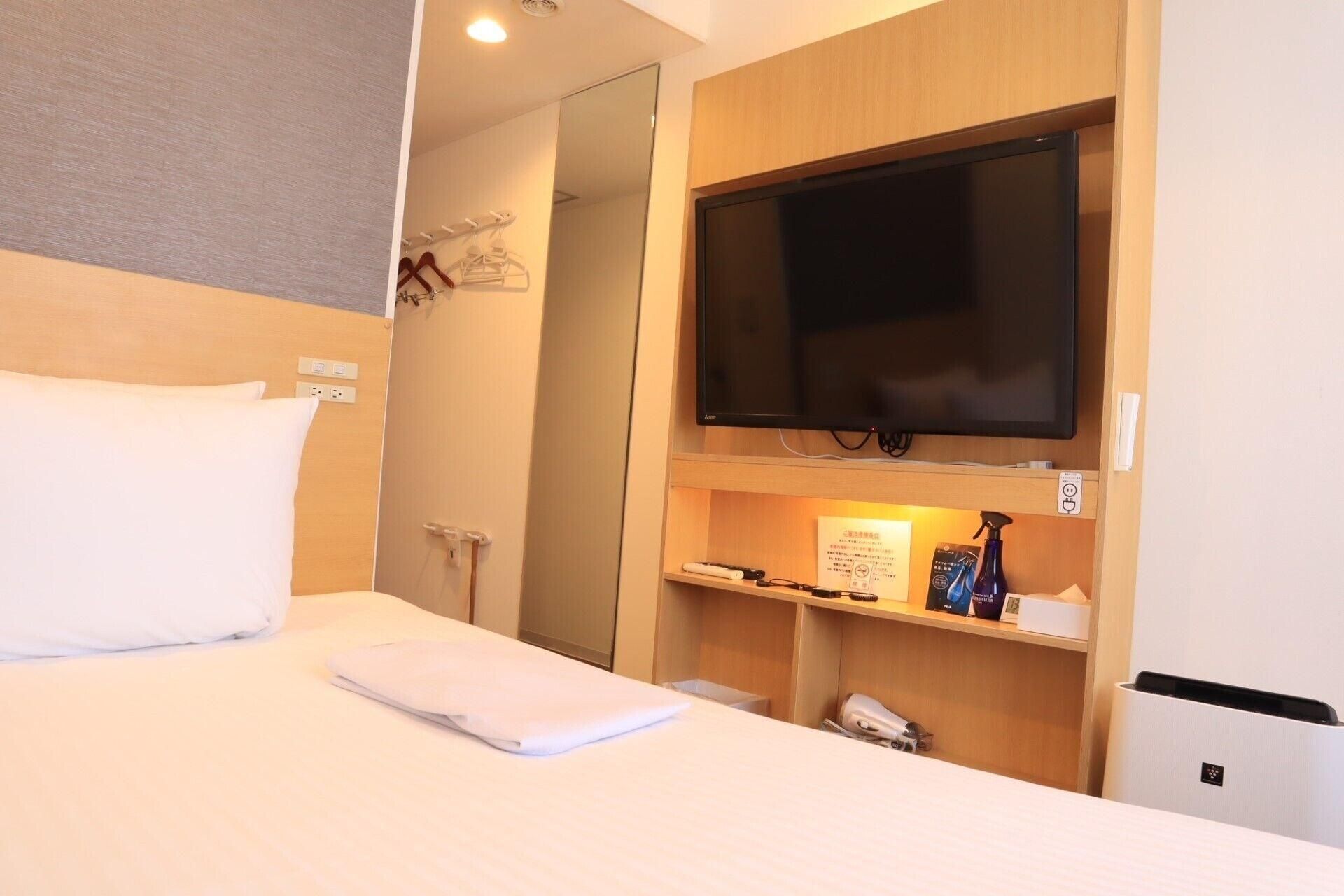 Habitación de hotel con una cama, un televisor montado en la pared, un estante con objetos y una puerta que da a otra habitación.