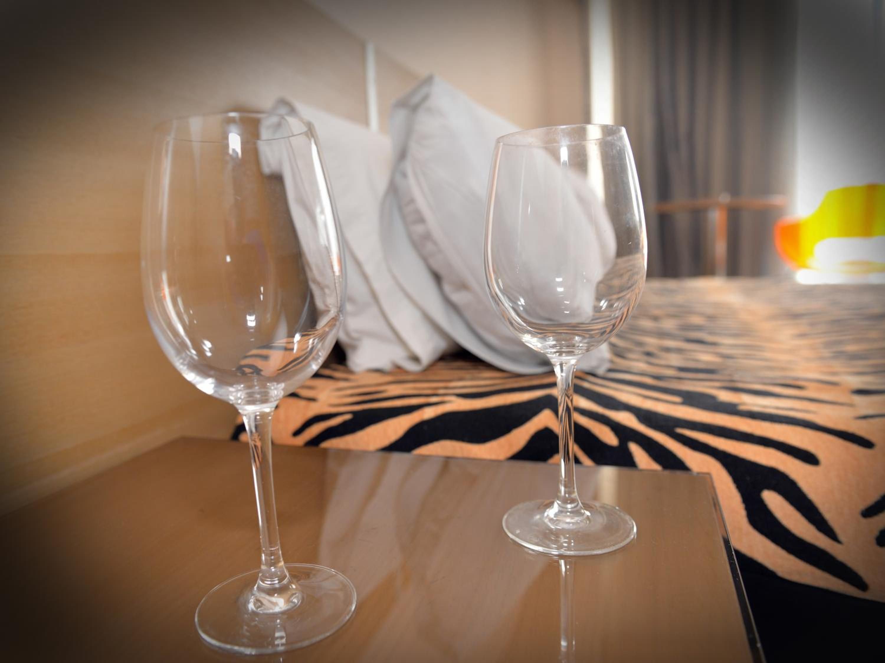 Two empty wine glasses on a table in a hotel room with a bed in the background.