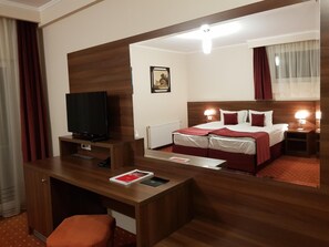 A hotel room with a wooden desk, a television, a bed with red bedding, and a nightstand with a lamp.