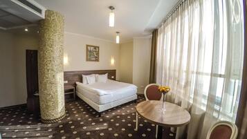 Ein Hotelzimmer mit einem Bett, einem runden Tisch mit einer Vase, einem Stuhl, einer großen Säule und einem Gemälde an der Wand.