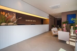 Una recepción con un letrero que dice 'BROWN-DOT HOTEL', una zona de estar con sillas y plantas en macetas.