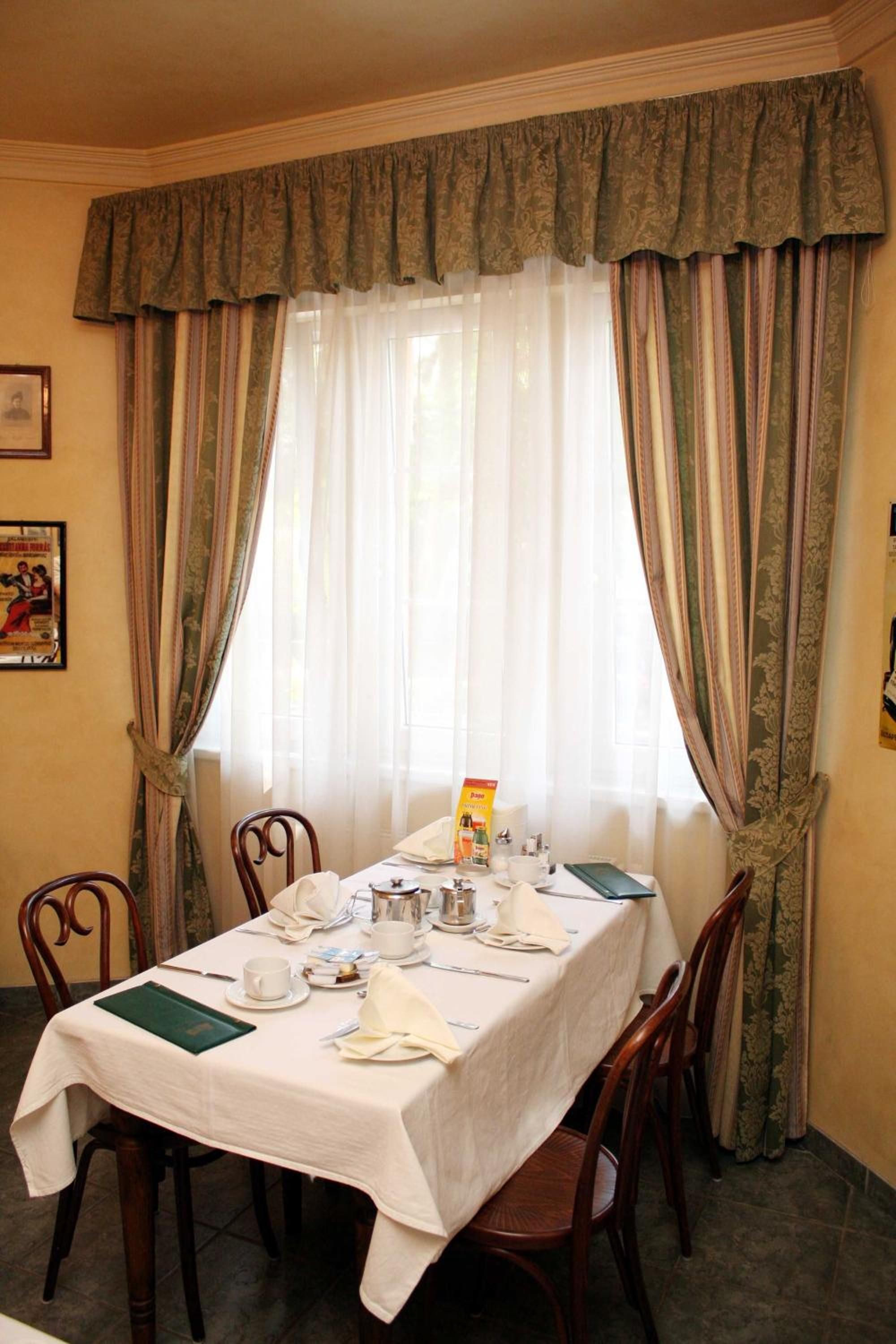 A dining room with a table set for two, featuring a white tablecloth, chairs, and a window with sheer curtains.