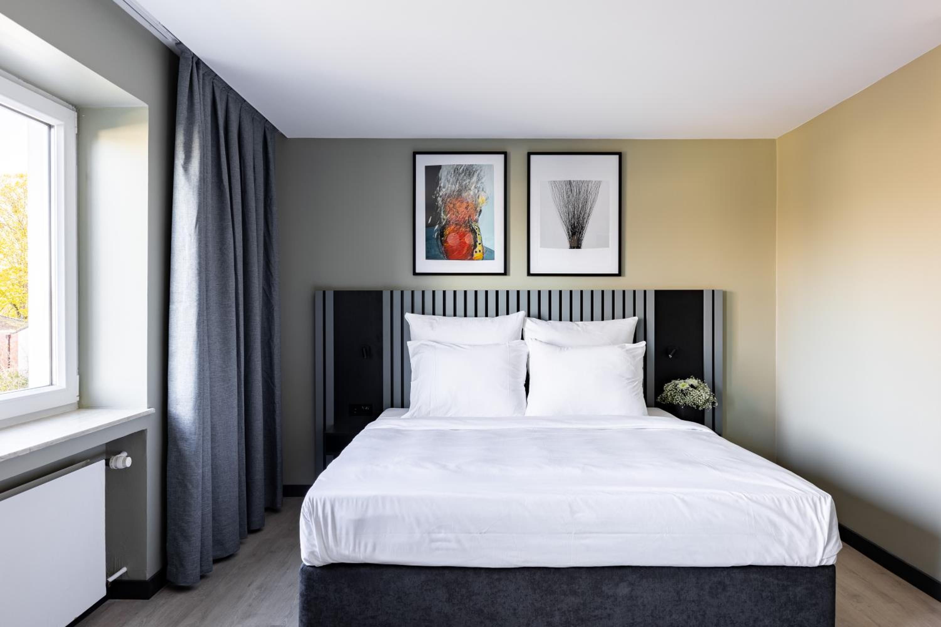 Une chambre d’hôtel moderne avec un grand lit, des rideaux gris et deux œuvres d’art encadrées accrochées au mur.