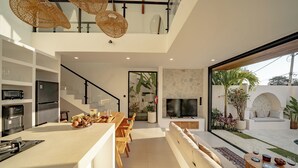 A modern kitchen with a central island, a dining area, and a living space with a TV and seating. - VIlla Ezra Azul (Uluwatu)
