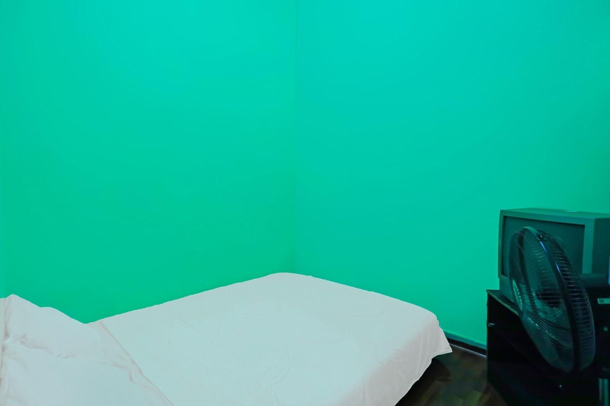 A room with a green wall, a bed with a white sheet, and a black fan on a stand.