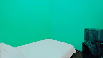 A room with a green wall, a bed with a white sheet, and a black fan on a stand.