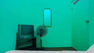 A room with a bed, a desk with a computer monitor, a standing fan, and a door.