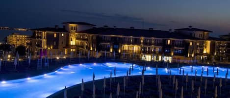 A hotel with a swimming pool, lounge chairs, and a well-lit building facade.
