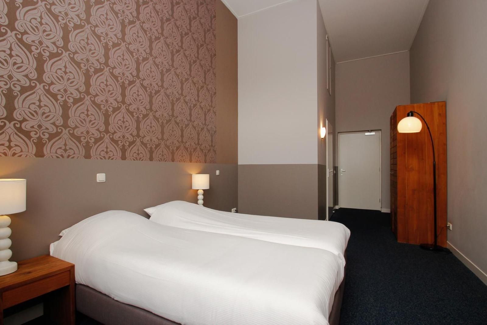 A hotel room with two beds, a patterned wallpaper, a wooden wardrobe, and a nightstand with a lamp.