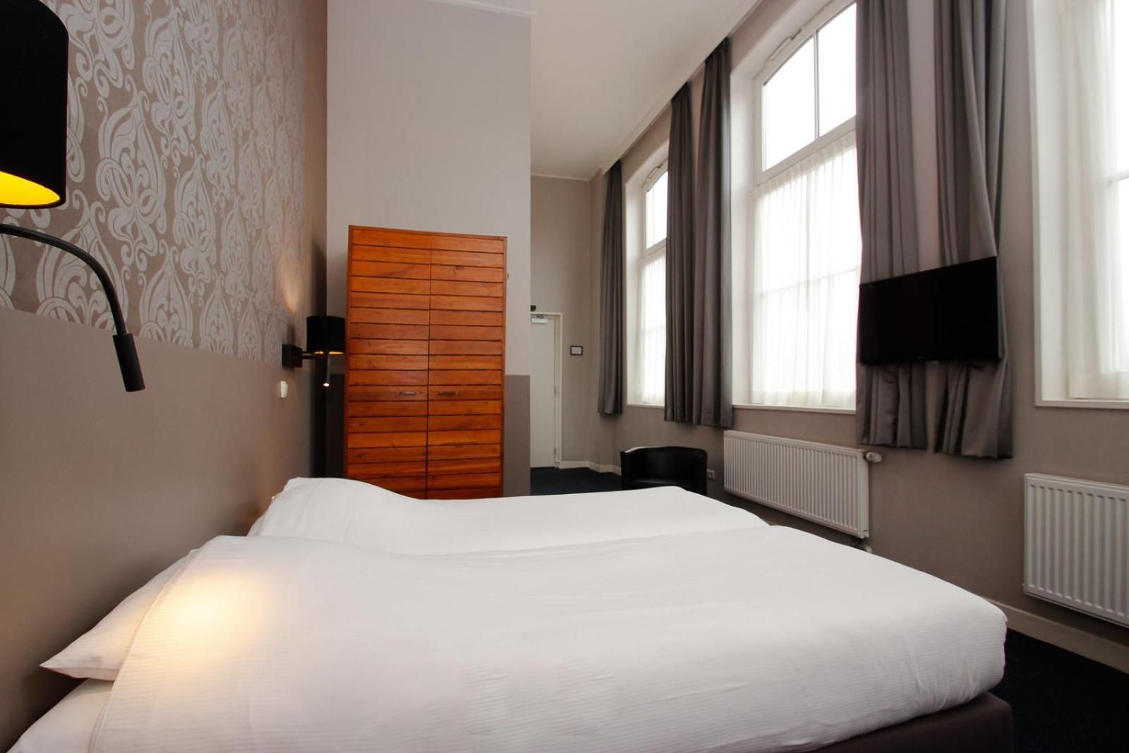 Ein Hotelzimmer mit einem Bett, einem Kleiderschrank, einem Schreibtisch, einem Stuhl, einem Fernseher und einem Fenster mit Vorhängen.