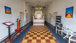 Un hall de hotel con mostrador de recepción, mesa de recepción, zona de espera y un rincón de lectura con una silla, una pequeña mesa y una estantería.