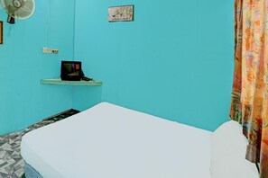 Um quarto com uma cama, uma pequena estante com uma televisão, um ventilador fixado na parede e uma cortina.