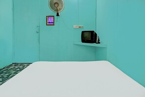 A room with teal walls, a white bed, a small shelf with a black television, a wall-mounted fan, and a purple light. - Spot On 92167 Jodoh Stay Syariah (Batam)