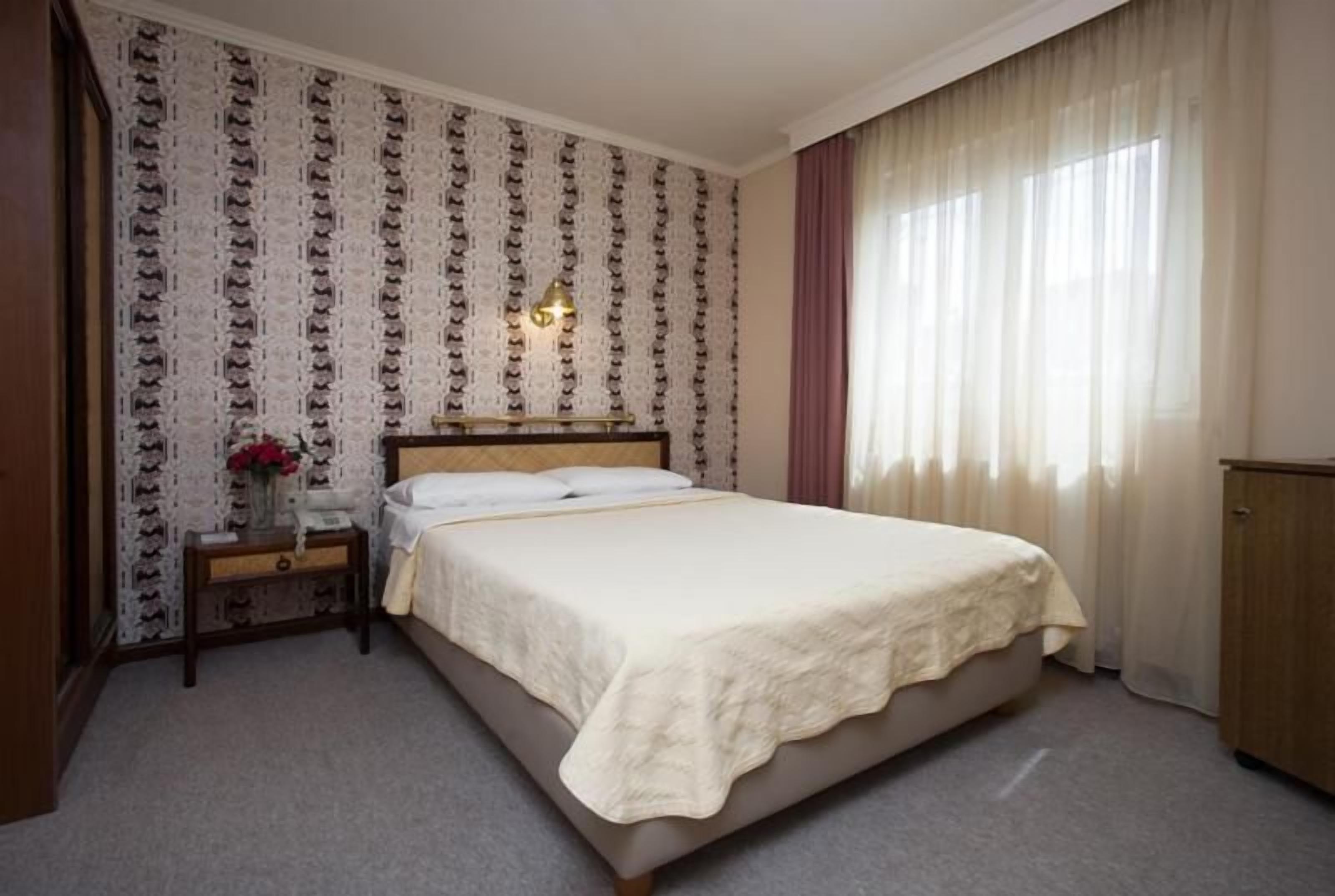 A hotel room with a bed, a nightstand, a lamp, a window with curtains, and a wall with a patterned wallpaper.
