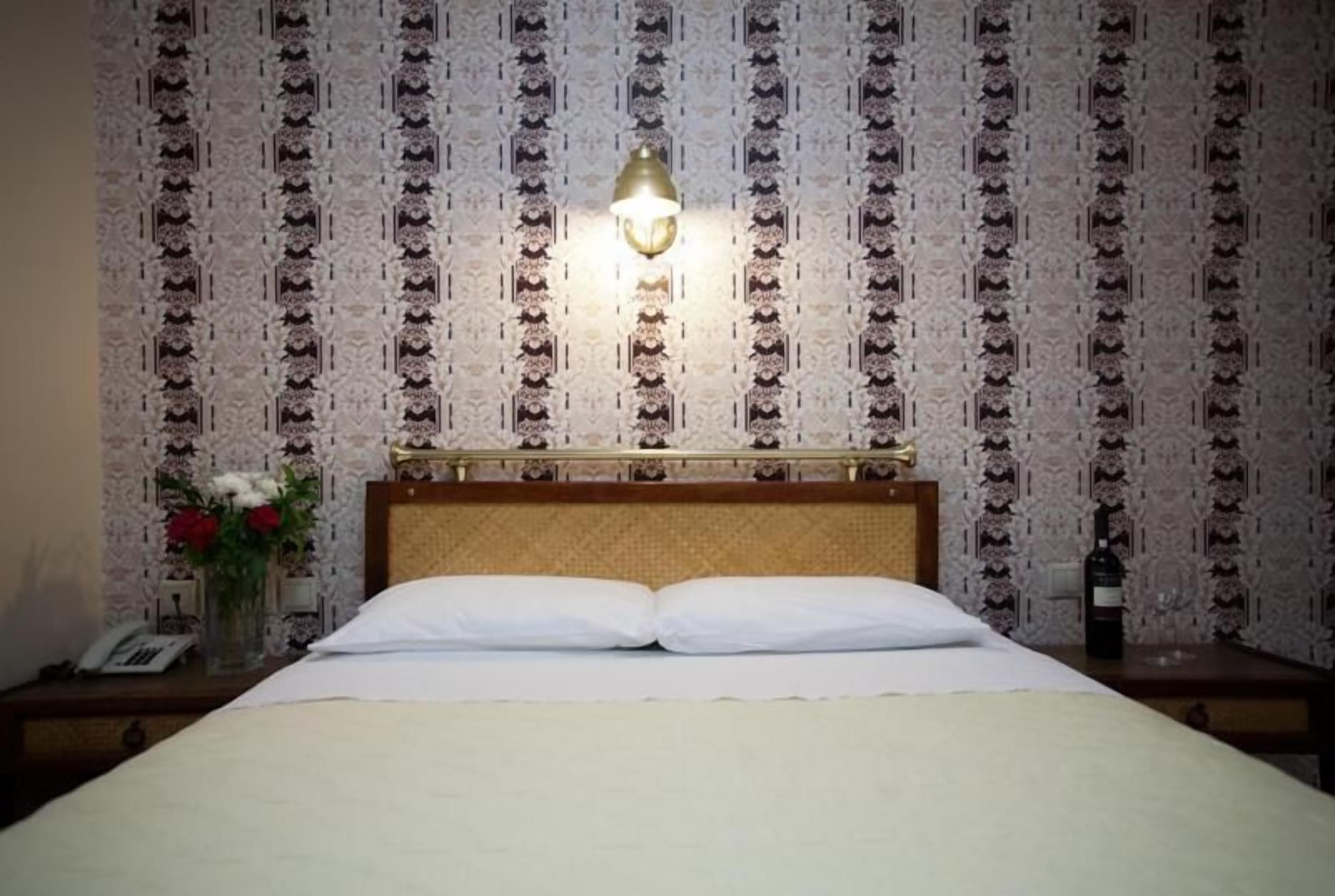 A hotel room with a bed, a nightstand, a lamp, and a wall with patterned wallpaper.