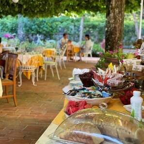An outdoor dining area with tables and chairs, a buffet with various food items, and a tree in the background. - Hotel Bellonda (Forte dei Marmi)