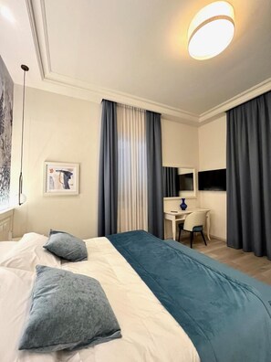 A hotel room with a bed, a desk, and a chair. There is a window with curtains, a wall-mounted TV, and a framed picture on the wall. - Hotel Bellonda (Forte dei Marmi)