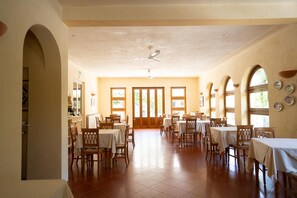 A spacious dining area with multiple tables and chairs, large windows, and a ceiling fan.