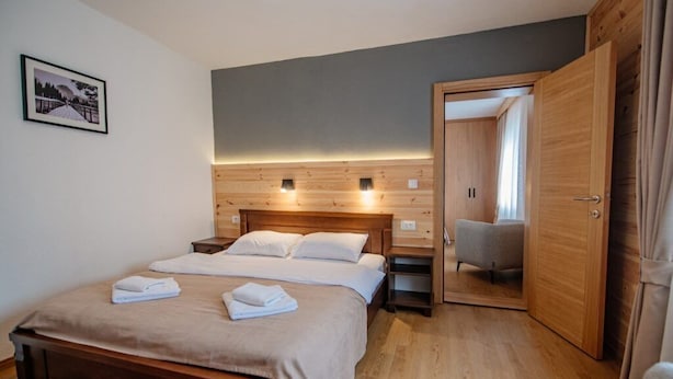 Quarto com cama de madeira, mesas de cabeceira, um criado-mudo, um espelho e uma porta que dá para outro espaço.
