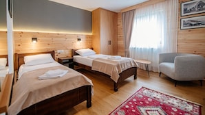 Quarto de hotel com duas camas, uma mesinha, uma cadeira e uma janela grande com cortinas.