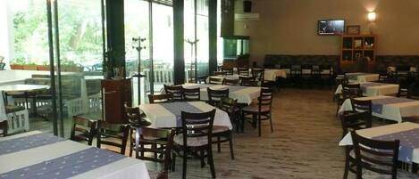 A restaurant with multiple tables and chairs, large windows, and a television on the wall.