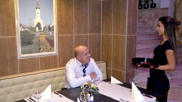 A restaurant setting with a dining table set for two, a waiter standing by, and a framed picture of a building on the wall. - Hotel Minerva (Mosonmagyarovar)