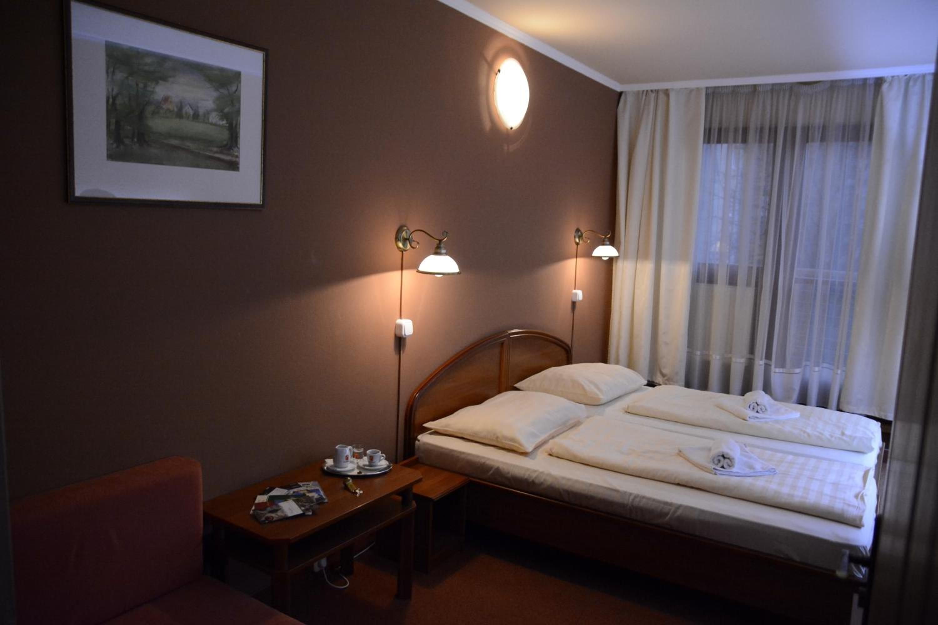 Ein Hotelzimmer mit einem Bett, Nachttischlampen, einem kleinen Tisch mit Telefon und Tasse, einem Sessel und einem Fenster mit Vorhängen.