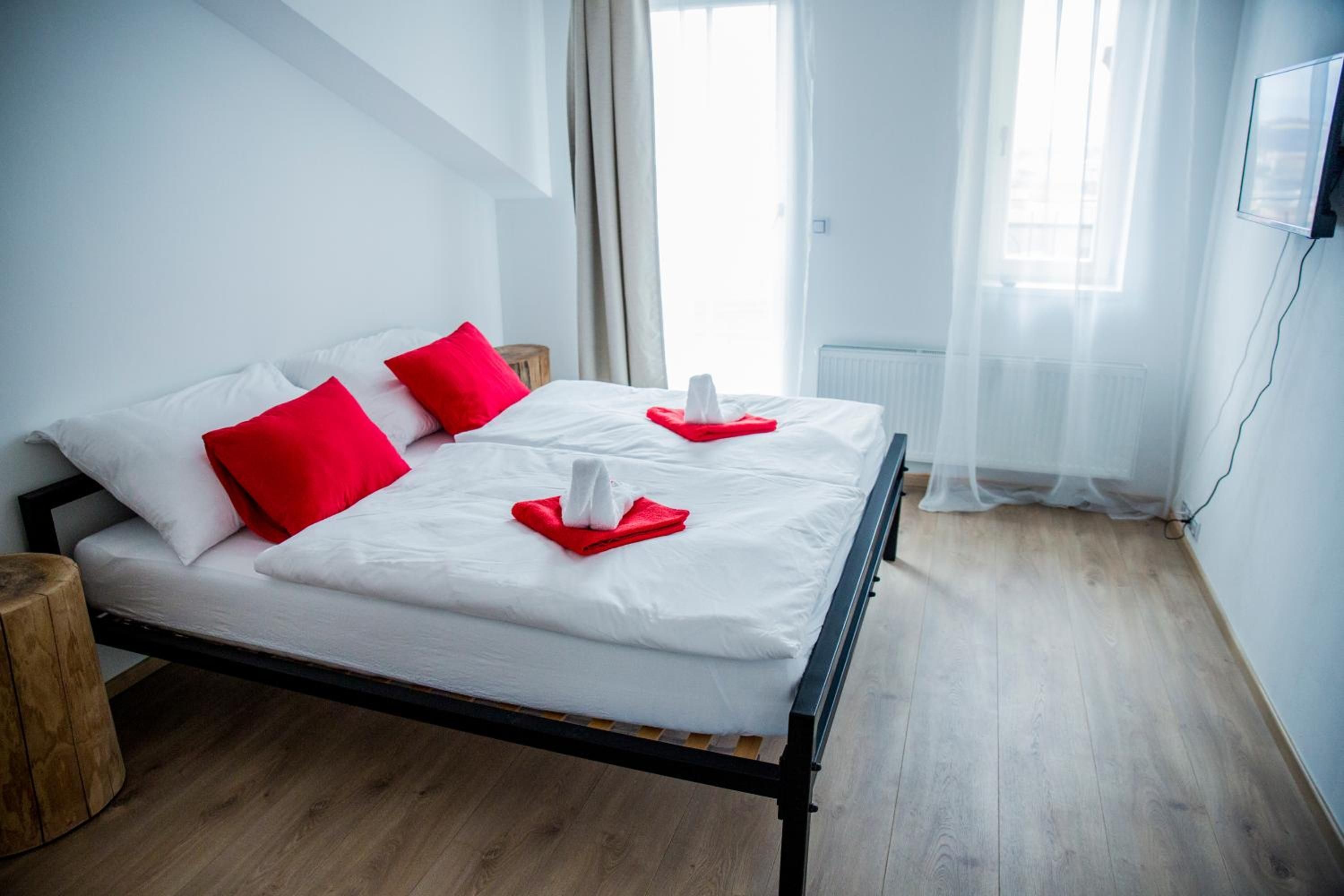 Una cama bien hecha con ropa blanca y almohadas rojas, una mesita de noche de madera y vistas de la ciudad a través de cortinas traslúcidas.