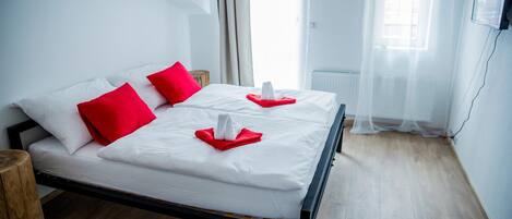 Una cama bien hecha con ropa blanca y almohadas rojas, una mesita de noche de madera y vistas de la ciudad a través de cortinas traslúcidas.