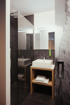 A modern bathroom with a glass shower enclosure, a white sink, and a wooden shelf with towels. - Hotel Leonard (Nova Ponente)