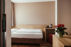 A hotel room with a bed, a small bedside table, a red ottoman, and a potted plant.