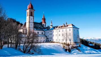 Ein historisches Gebäude mit einem markanten Turm und zahlreichen Spitzbögen, umgeben von verschneiter Landschaft und Bäumen.