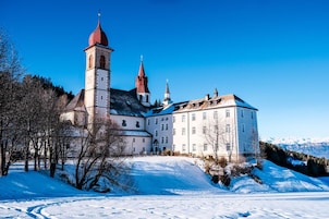 A historic building with a prominent tower and multiple spires, surrounded by snow-covered landscape and trees.
