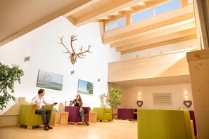 Un vaste espace intérieur agrémenté de poutres en bois, d’une grande sculpture représentant une tête de cerf fixée au mur, et de plusieurs coins salon avec des coussins aux couleurs vives.