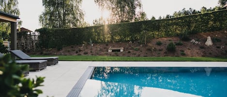 A swimming pool with a tiled deck, surrounded by a landscaped garden with a hedge and trees.