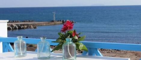 A white table with three glass bottles and a potted plant with red flowers, overlooking a beach and the sea.