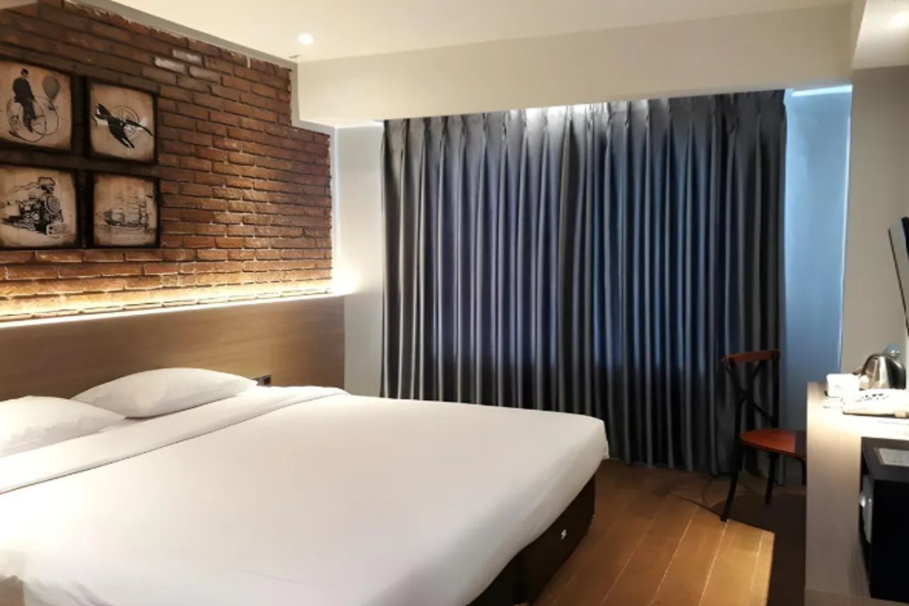 Une chambre d’hôtel avec un lit, un bureau, une chaise, une fenêtre avec des rideaux et un mur en briques orné de tableaux encadrés.