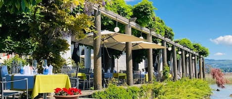 An outdoor dining area with tables and chairs, a river in the background, and a pergola structure.