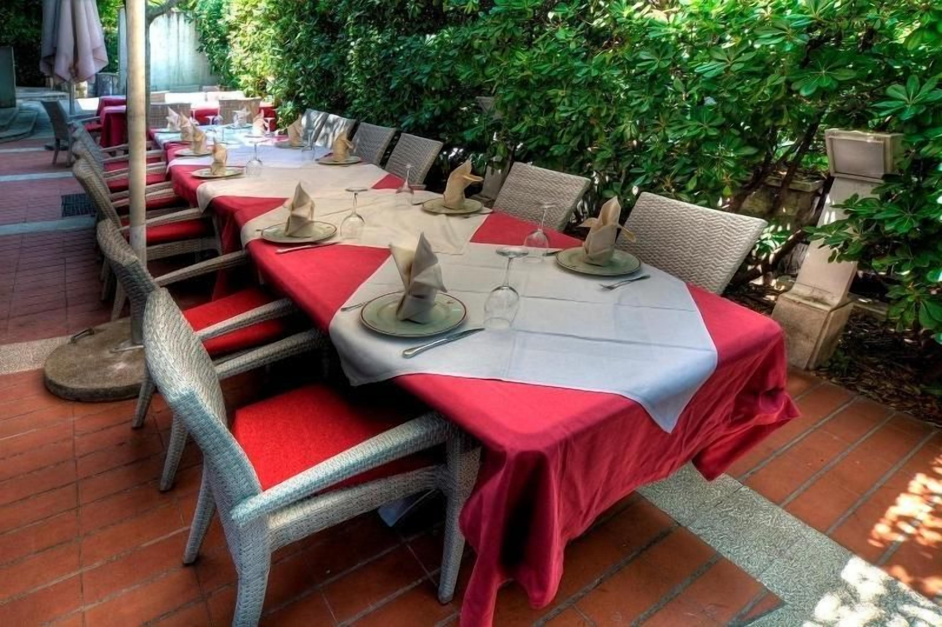 Un'area pranzo all'aperto con tavoli apparecchiati per due, sedie in vimini e pavimento in mattoni.