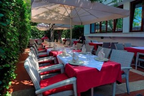 An outdoor dining area with tables set for two, covered by umbrellas, and surrounded by greenery. - Hotel Consul (Split)