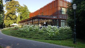 Edificio moderno con área de estar techada al aire libre, rodeado de abundante vegetación y flores blancas.
