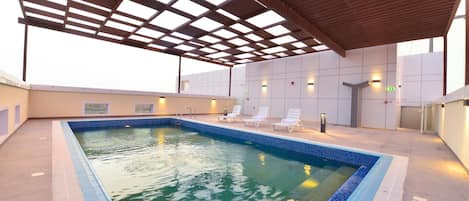 A rooftop pool with a tiled edge, surrounded by lounge chairs and a covered area with a wooden roof.