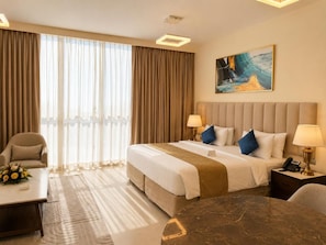 A hotel room with a bed, two bedside tables, a desk, a chair, a large window with curtains, and a painting on the wall. - Saray Deluxe Hotel Apartments (Abu Dhabi)