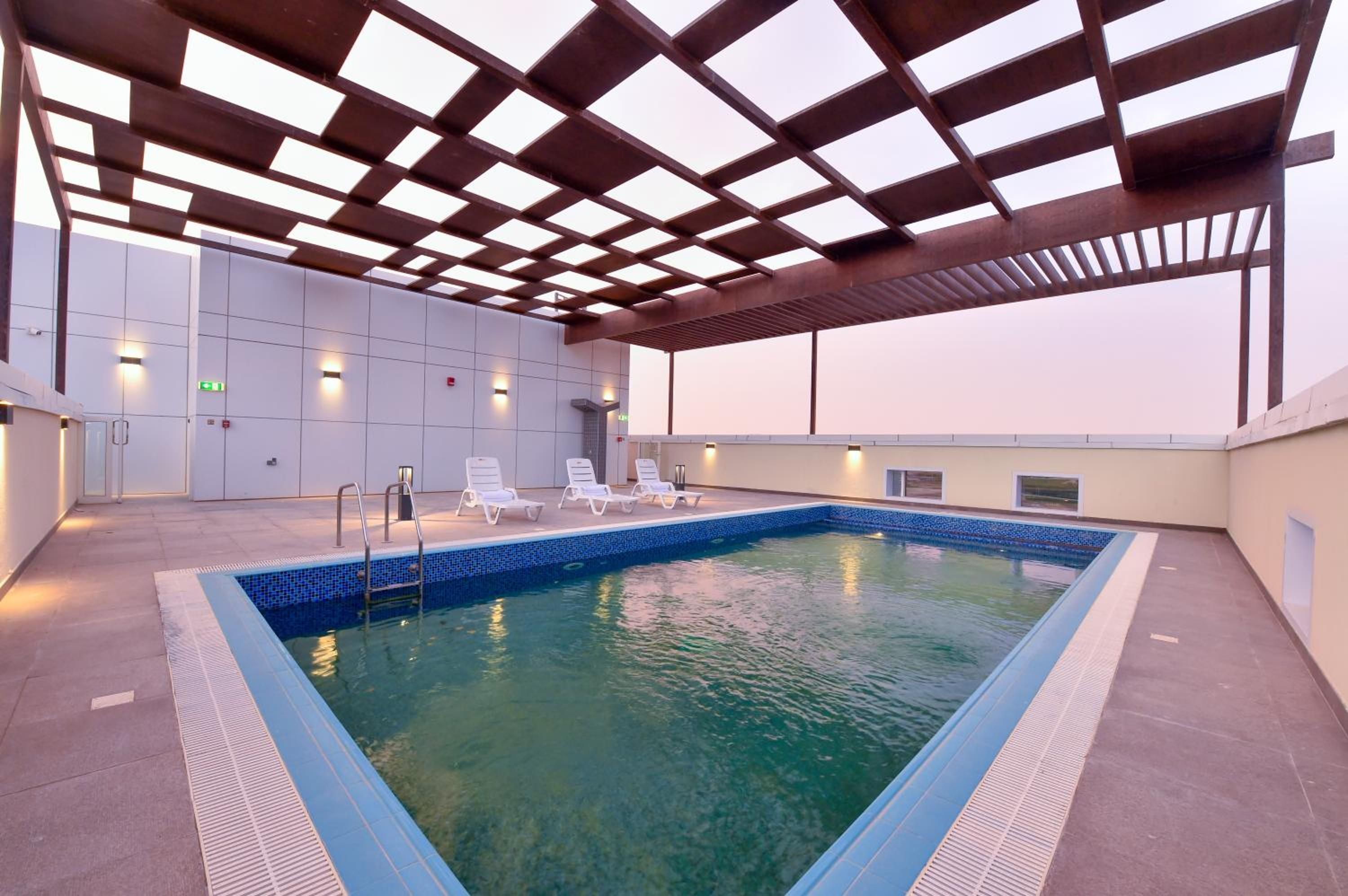 A rooftop swimming pool with a clear blue water feature, surrounded by lounge chairs and a covered area with a metal framework.