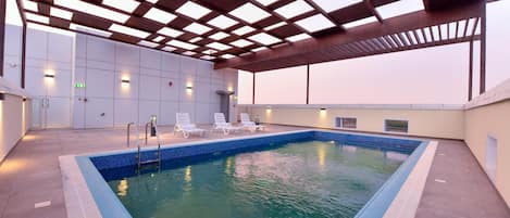 A rooftop swimming pool with a clear blue water feature, surrounded by lounge chairs and a covered area with a metal framework.