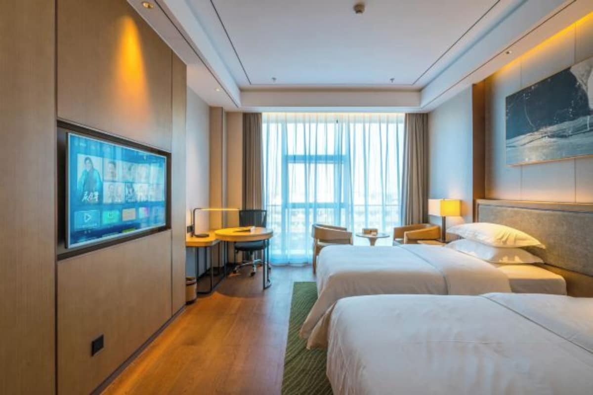 A hotel room with two beds, a TV, a desk, and a large window.