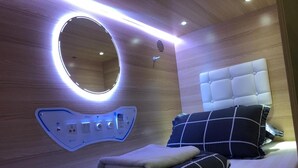 A modern bedroom with a circular mirror, a bed with a tufted headboard, and a bedside table with a power strip. - Capsulepod@aljunied (Singapore)