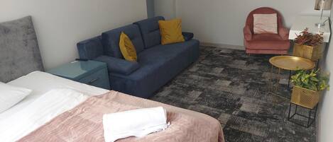 Una habitación con cama, un sofá azul con almohadones amarillos, un sillón rosa, una mesita redonda con una planta y un cabecero gris.